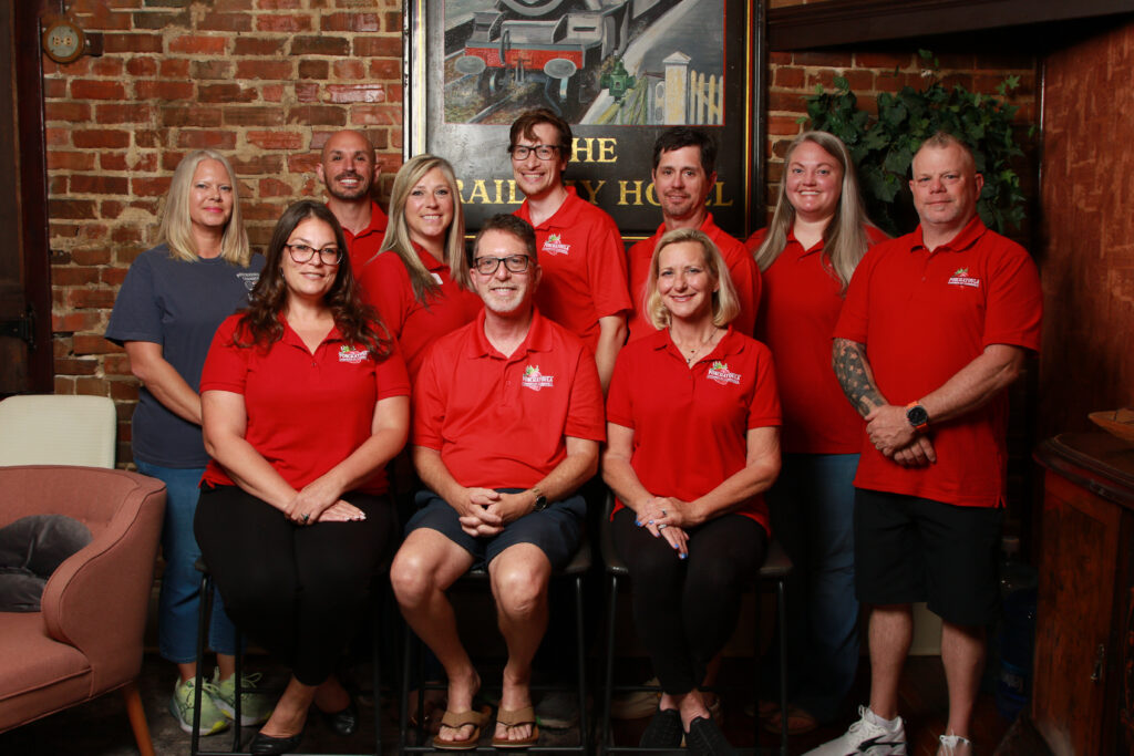 Group of ten staff members in red polo shirts posing for a group photo indoors against a brick wall.