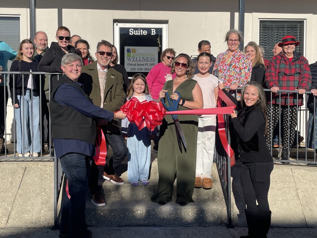 Group of people at a ribbon-cutting ceremony in front of a building labeled 'Suite B', with a large red bow and oversized scissors visible in the foreground.