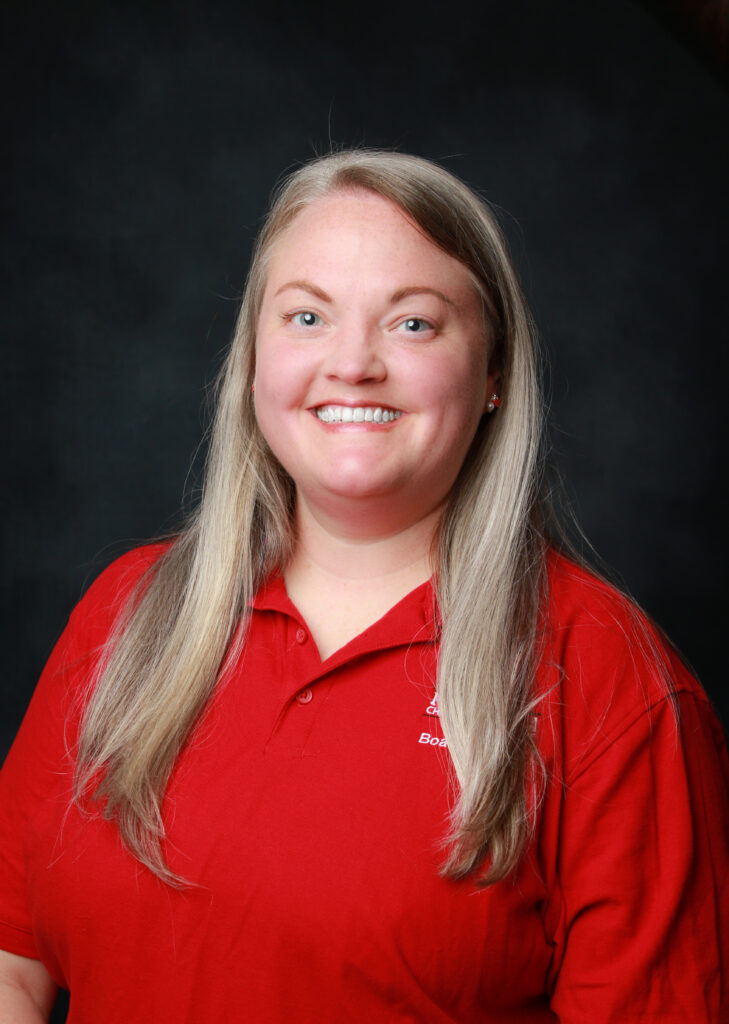 Smiling woman with long blond hair wearing a red polo shirt against a dark backdrop.