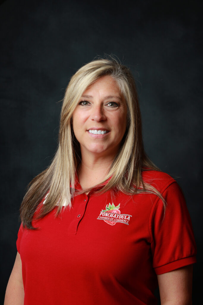 Head-and-shoulders portrait of a smiling woman with blonde hair wearing a red polo shirt against a dark background, logo on shirt.